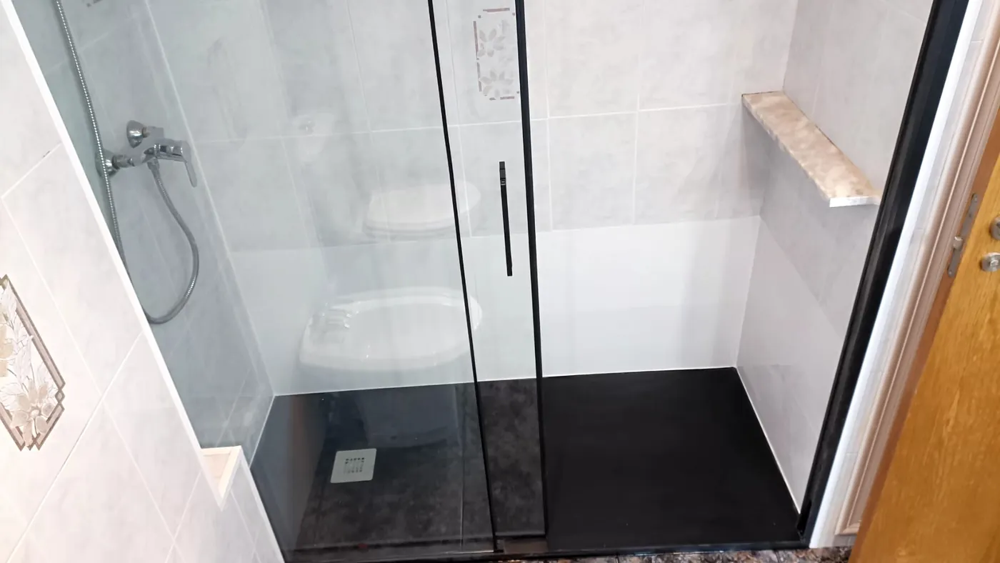 Después: plato de ducha negro tipo pizarra a ras de suelo con mampara de perfil negro y grifería monomando, en el mismo baño