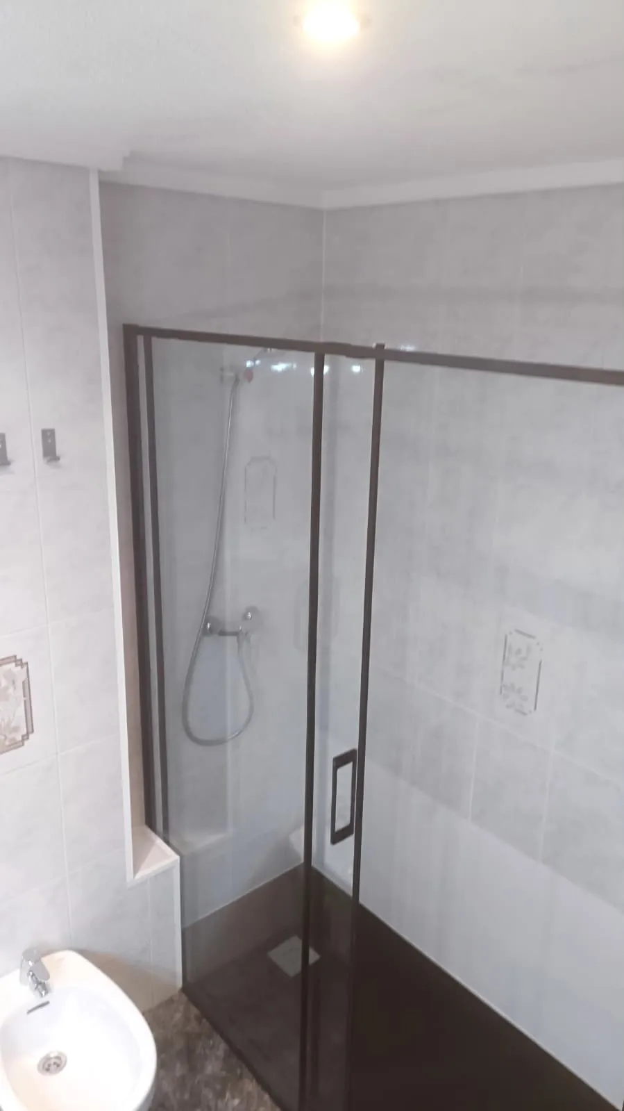 Ducha instalada donde había una bañera, con plato oscuro antideslizante, mampara corredera de dos hojas con perfil marrón y alicatado gris
