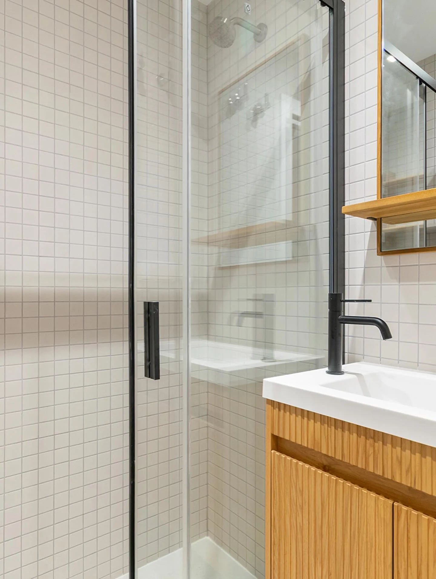 Ducha con gresite blanco, mampara de perfil negro y mueble de roble ranurado con grifería negra en un baño de Alicante